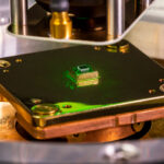 a small square chip sits on a metallic microscope stand with green laser light bouncing off of it in places
